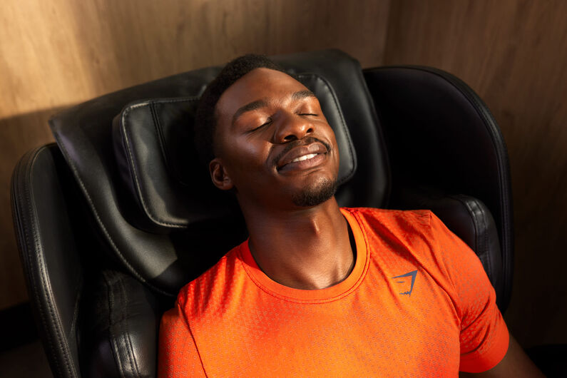 A man relaxes in a massage chair at a Basic-Fit gym, eyes closed and smiling after a workout. He wears an orange sports shirt in a calm recovery area.