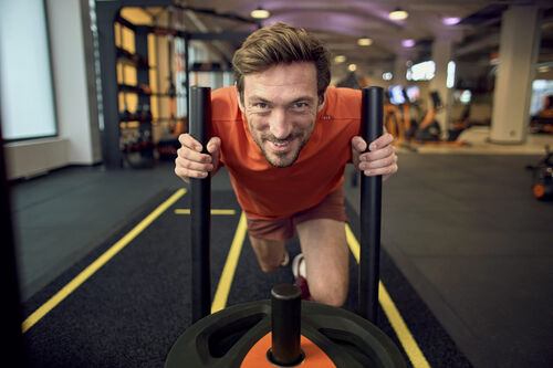 A man pushes a weighted sled during a workout in a Basic-Fit gym, smiling as he trains in the functional training area.